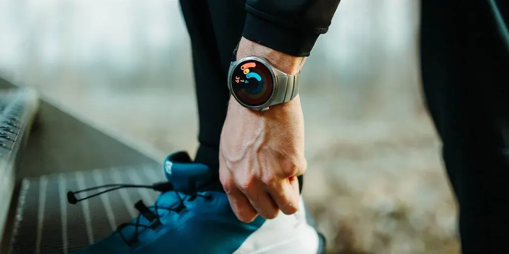 Eine Hand mit einer silbernen Smartwatch am Handgelenk bindet einen blauen Sportschuh. Die Uhr zeigt bunte Aktivitätsringe an.