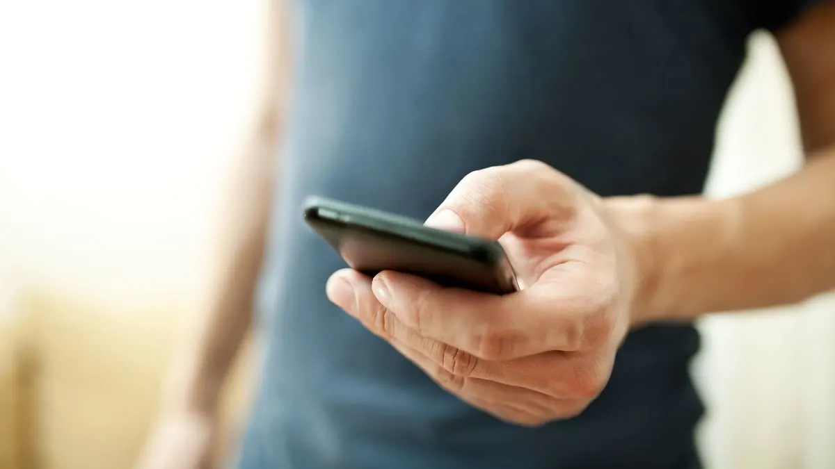 Nahaufnahme einer Hand, die ein schwarzes Smartphone hält. Eine Person in einem dunkelblauen Hemd steht vor einem verschwommenen hellen Hintergrund.
