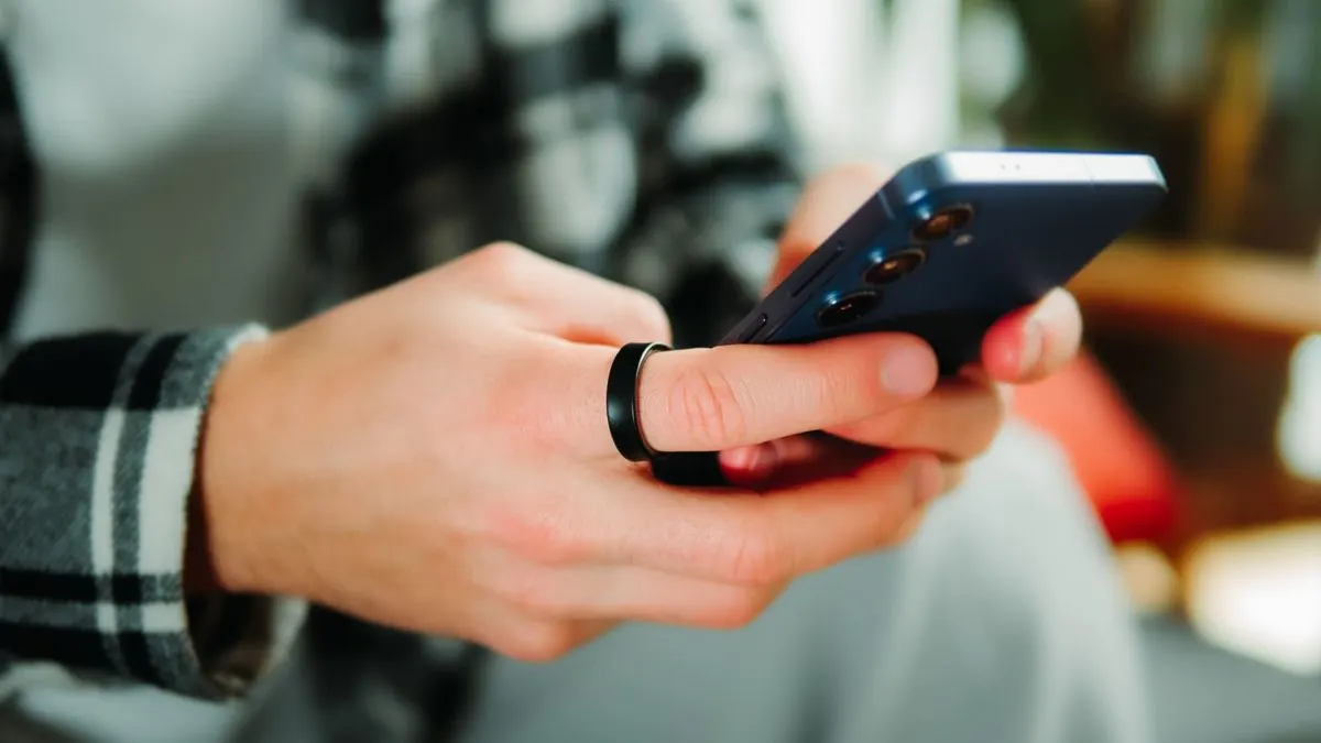 Person mit kariertem Hemd hält ein Smartphone in der Hand, am Finger ein schwarzer Smart-Ring.