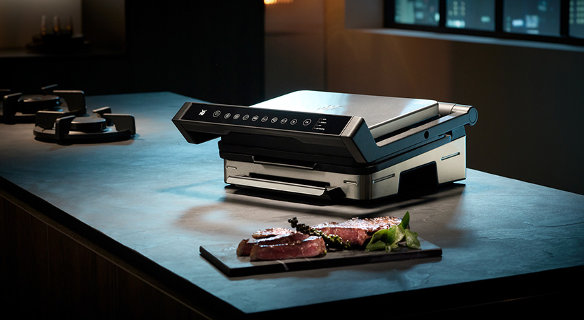 Ein elektrischer Grill steht auf einer Küchentheke, daneben ein Teller mit Fleisch und Kräutern. Im Hintergrund ist ein Kochfeld.