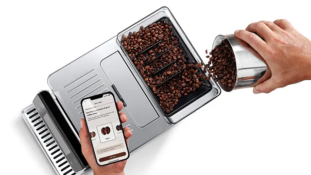 Kaffeebohnen werden in eine silberne Kaffeemaschine gefüllt, während ein Mobiltelefon gehalten wird.