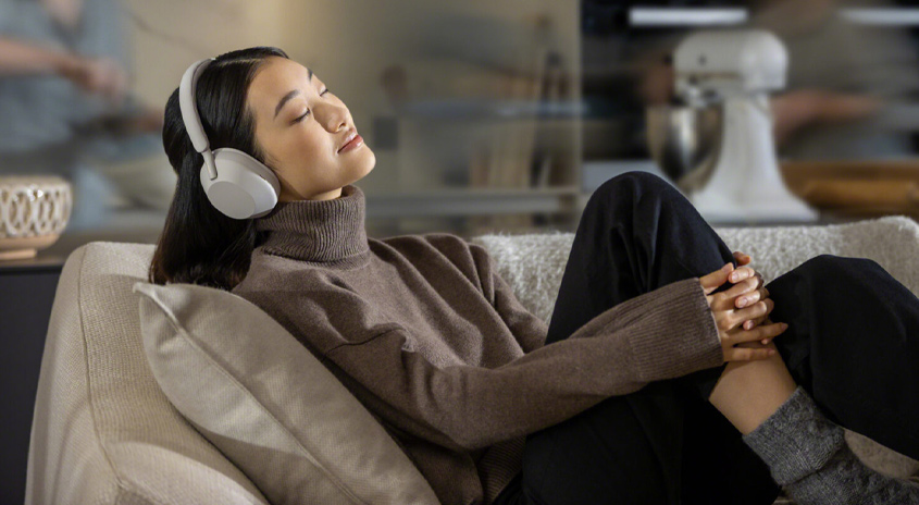 Eine Frau mit Kopfhörern liegt auf einer Couch. Sie trägt einen braunen Rollkragenpullover und schwarze Hosen. Im Hintergrund ein Küchengerät.