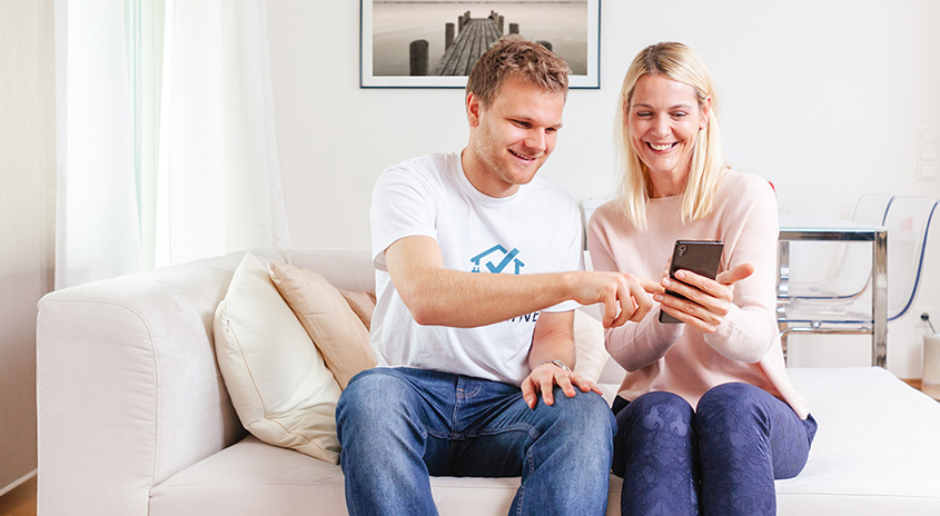 Ein Paar sitzt auf einem weißen Sofa und schaut auf ein Smartphone. Der Mann zeigt auf den Bildschirm.