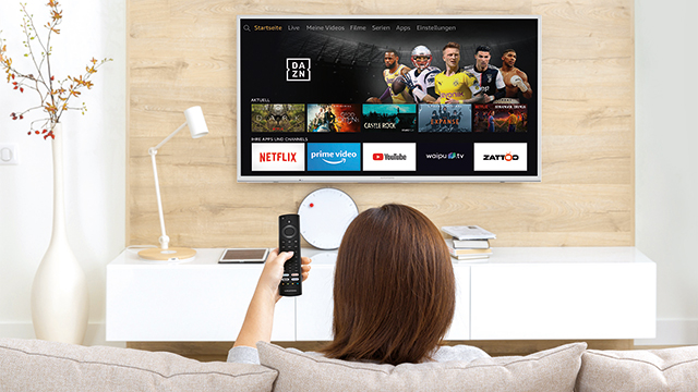 Eine Frau sitzt auf einem Sofa und hält eine Fernbedienung vor einem Fernseher mit Streaming-Apps.