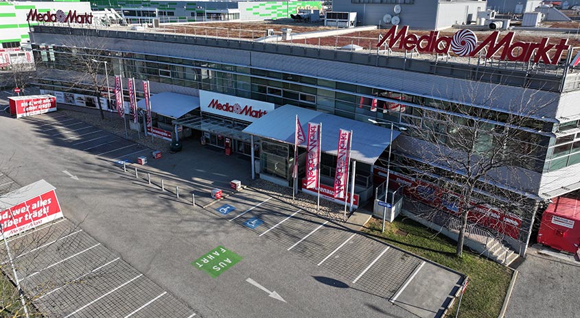 Ansicht eines MediaMarkt-Gebäudes mit Parkplatz. Das Gebäude hat eine Glasfassade und ein rotes Logo. Parkplätze mit Markierungen sind sichtbar.
