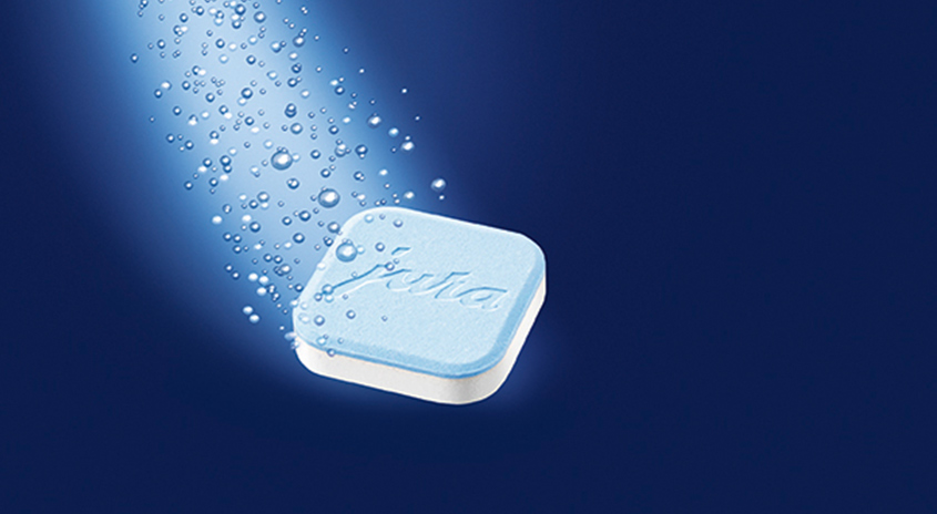Eine hellblaue, quadratische Reinigungstablette mit weißer Unterseite schwebt vor einem dunkelblauen Hintergrund mit Wassertropfen.