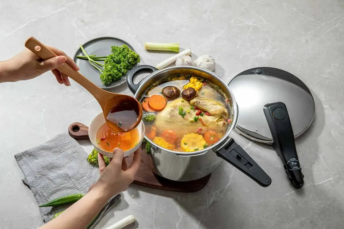 Zwei Hände schöpfen Hühner-Gemüse-Suppe aus einem Schnellkochtopf in eine kleine Schüssel, dahinter liegen der Deckel und rohe Zutaten.