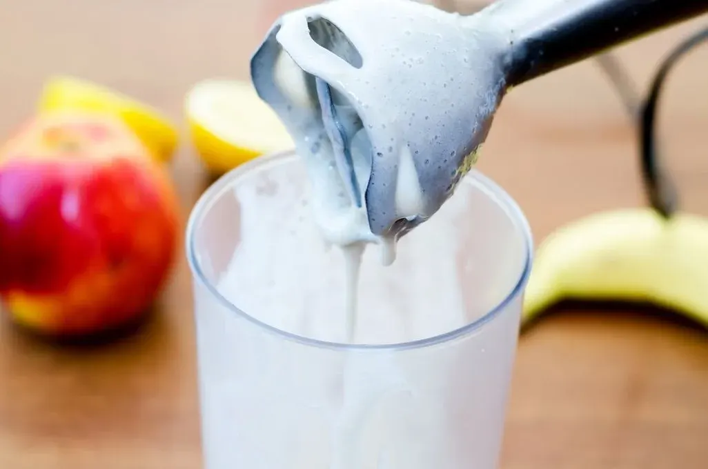 Stabmixer mit Behälter in dem sich eine weiße Masse befindet, im Hintergrund sieht man Obst