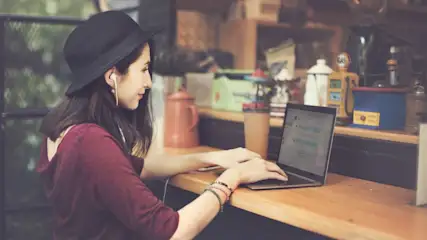 Frau mit schwarzem Hut arbeitet konzentriert am Laptop in gemütlichem Café.
