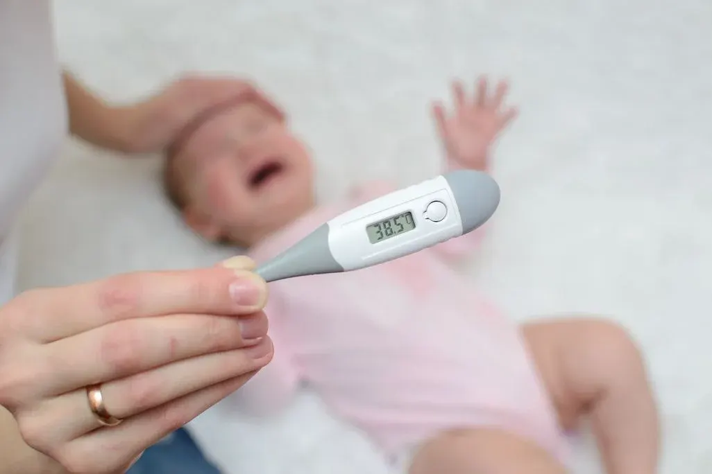 Baby weint im Hintergrund, Frau hält einen Fieberthermometer, der 38,5 Grad anzeigt.