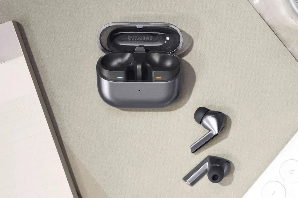 Silberfarbene Samsung Galaxy Buds3 liegen auf einer Kartonfläche vor ihrem geöffneten Ladecase. Im Anschnitt sind ein Block und ein Telefon zu sehen.