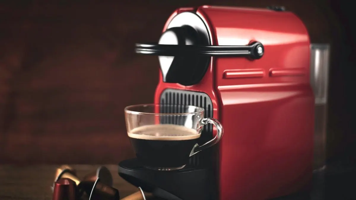 Rote Nespresso Kaffeemaschine mit einer klaren Glastasse voll dunklem Kaffee und Schaum. Bunte Kapseln liegen auf dem dunklen Holztisch vor einer Holzwand.