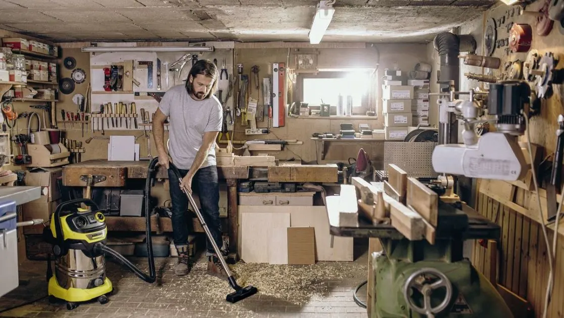 Mann saugt Holzspäne mit einem gelb-silbernen Staubsauger in einer voll ausgestatteten Werkstatt. Werkzeuge und Maschinen sind sichtbar.