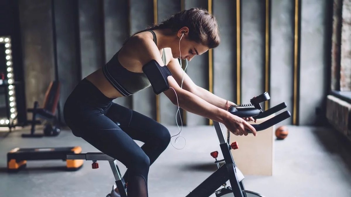 Eine Frau in schwarzer Sportkleidung trainiert am Ergometer und hört dabei Musik mit Kopfhörern.