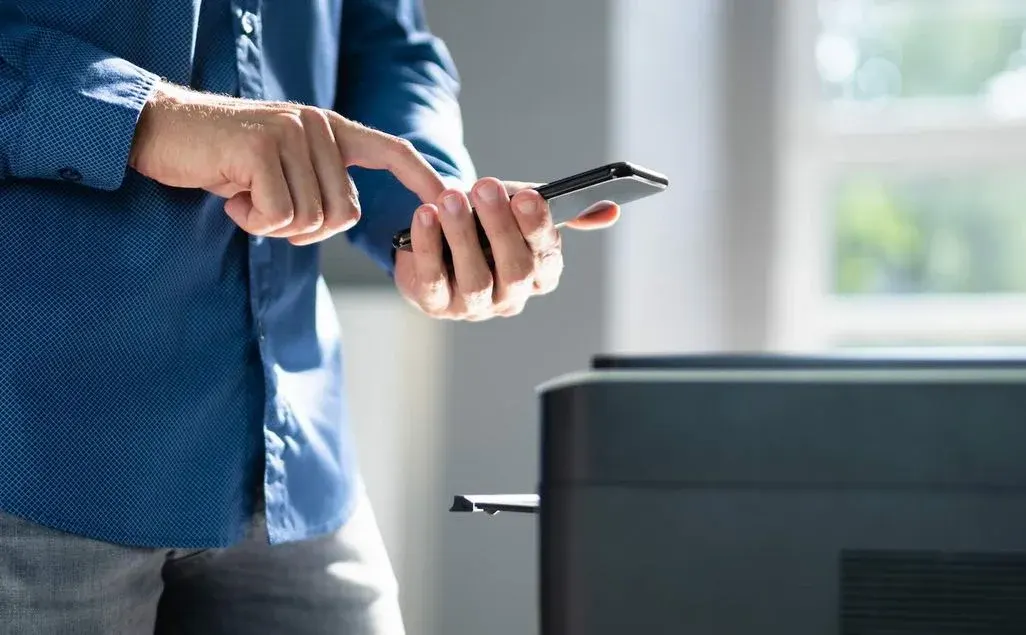 Person bedient Smartphone neben Drucker, Finger tippt auf Display.