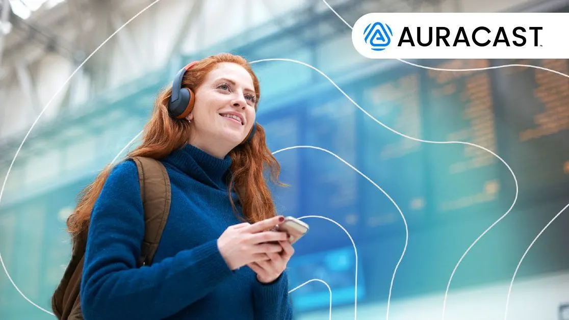 Person mit Kopfhörern und Rucksack steht vor Anzeigetafel, hält ein Smartphone. Oben rechts ist das Auracast-Logo.