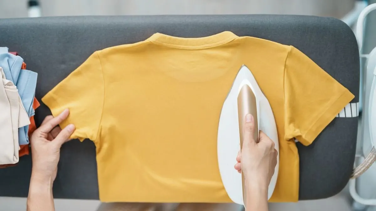 Person bügelt ein gelbes T-Shirt auf einem Bügelbrett. Daneben liegt ein Stapel gefalteter Kleidung.