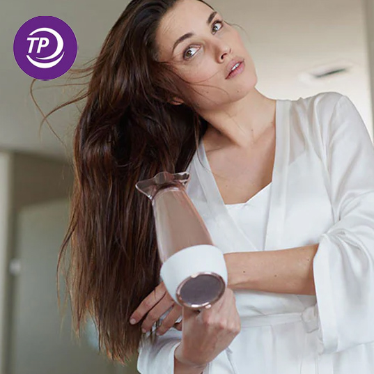 Eine Frau im Bademantel föhnt ihre Haare mit einem Föhn mit weißem und bronzefarbenem Gehäuse.