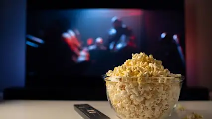 Eine Glasschale voller Popcorn und eine Fernbedienung liegen auf einem weißen Tisch vor einem unscharfen TV-Bild mit roten und blauen Lichtern.