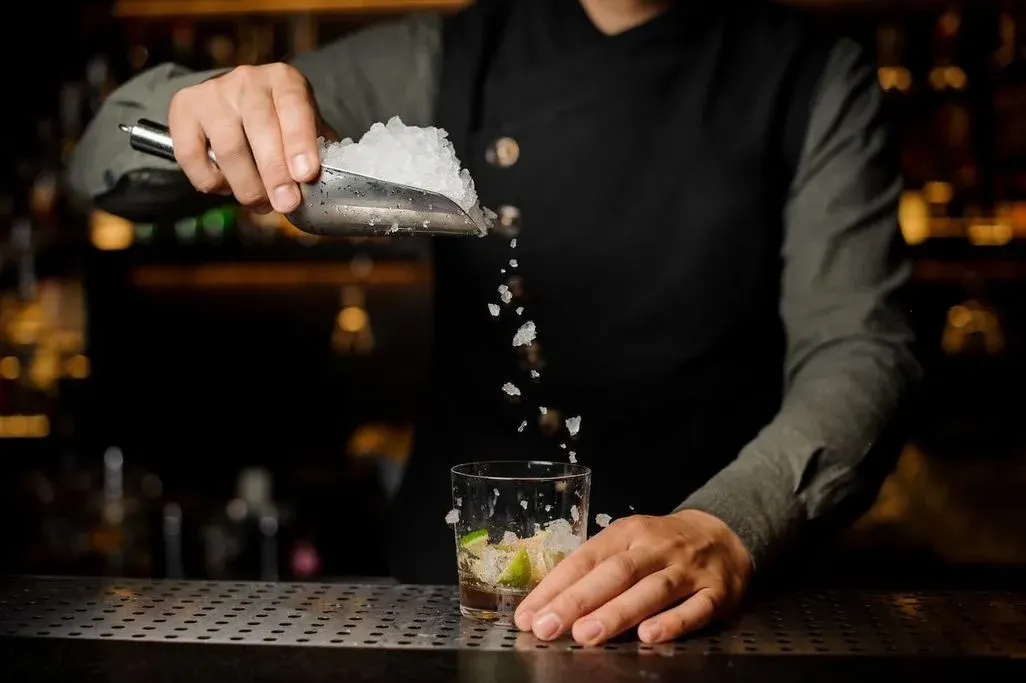 Barkeeper schüttet zerstoßenes Eis aus einer Schaufel in ein Glas mit Limettenstücken an der Bar.
