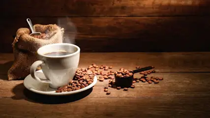 Weißer Kaffeebecher mit dampfendem Kaffee auf Untertasse, umgeben von Kaffeebohnen, neben einem Jutesack und Messlöffel auf Holztisch.