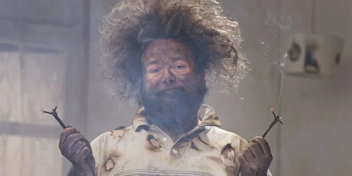 Mann mit zerzaustem Haar und verbranntem Hemd hält zwei durchgebrannte Kabelenden.