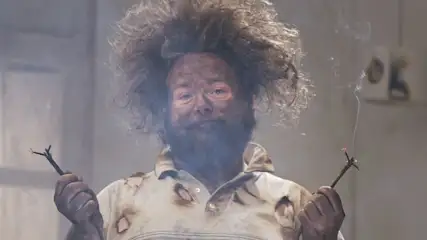 Mann mit zerzaustem Haar und verbranntem Hemd hält zwei durchgebrannte Kabelenden.