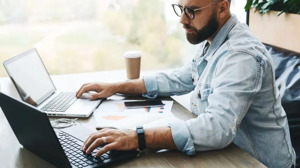 Mann sitzt am Schreibtisch und arbeitet an 2 Laptops gleichzeitig