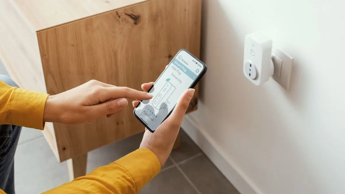 Hände halten ein Smartphone, das einen Smart Plug verbindet. Ein weißer Smart Plug steckt in einer Wandsteckdose neben einem Holzschrank.