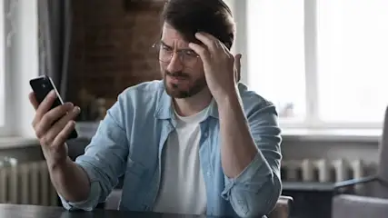 Ein Mann mit Brille und hellblauem Hemd hält ein schwarzes Smartphone, blickt besorgt darauf, Hand an Stirn. Sitzt an dunklem Tisch vor Ziegelwand und Fenster.