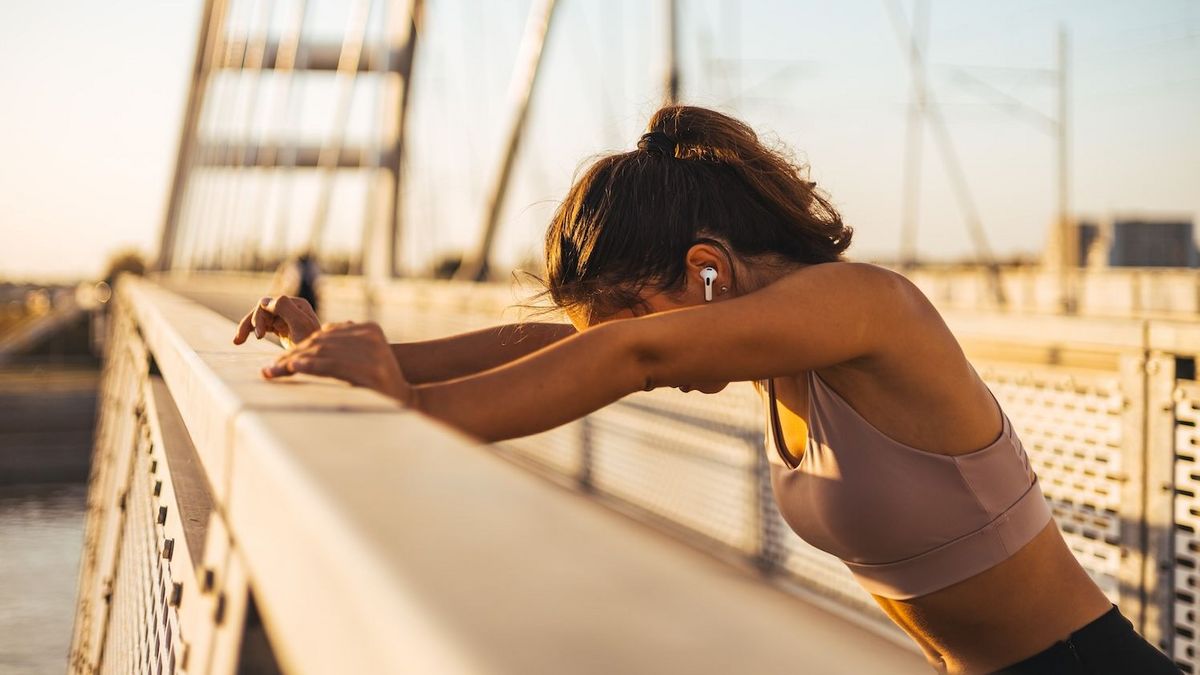 Person in Sportkleidung lehnt mit weißen Ohrhörern auf einer hellen Brüstung einer Brücke bei Sonnenuntergang über Wasser und Stadt.