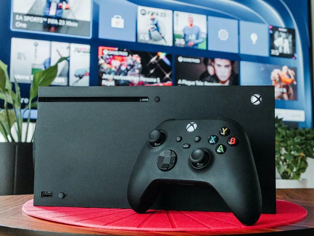 Eine schwarze Xbox-Konsole mit Controller steht vor einem Fernseher mit Spiele-Menü.
