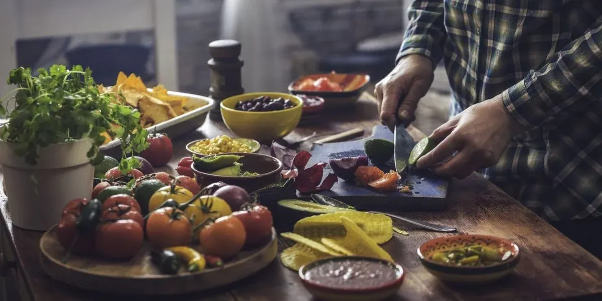 Hände in kariertem Hemd schneiden Limette auf Schneidebrett. Tisch voller frischer Tomaten, Paprika, Koriander, Avocado, Mais, Bohnen, Salsa und Tortilla-Chips.