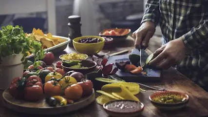 Hände in kariertem Hemd schneiden Limette auf Schneidebrett. Tisch voller frischer Tomaten, Paprika, Koriander, Avocado, Mais, Bohnen, Salsa und Tortilla-Chips.