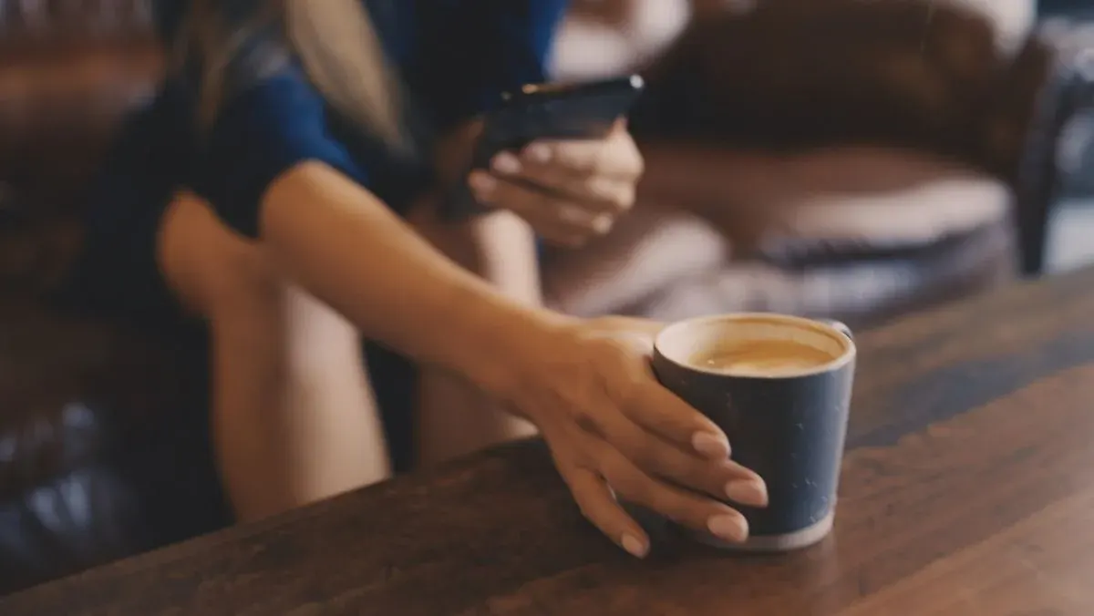 Person in blauem Kleid hält Smartphone, greift nach dunkelgrauer Kaffeetasse auf Holztisch. Braunes Sofa im Hintergrund.