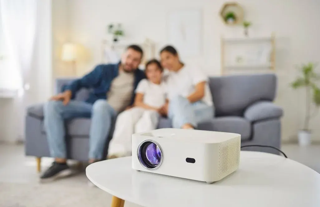 Weißer Beamer steht im Vordergrund auf einem Couchtisch, im Hintergrund sitzt eine lächelnde Familie unscharf auf dem Sofa im hellen Wohnzimmer.