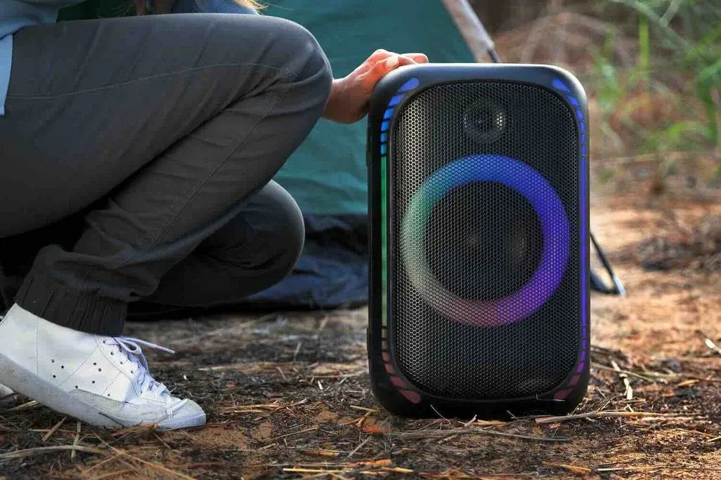 Eine Person mit grauer Hose und weißen Schuhen stellt vor einem grünen Zelt einen Outdoor-Lautsprecher ab.