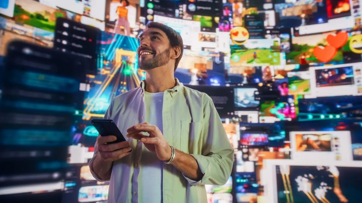 Lächelnder Mann mit Smartphone vor einer Wand aus zahlreichen digitalen Videos und Social-Media-Feeds in leuchtenden Farben.