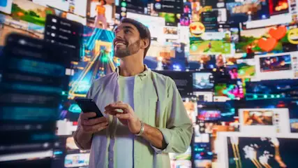 Lächelnder Mann mit Smartphone vor einer Wand aus zahlreichen digitalen Videos und Social-Media-Feeds in leuchtenden Farben.