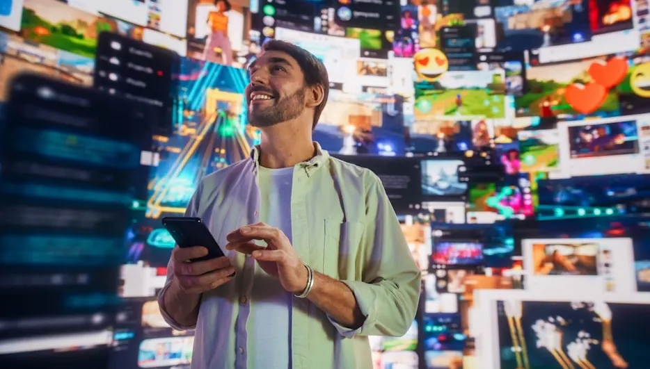 Lächelnder Mann mit Smartphone vor einer Wand aus zahlreichen digitalen Videos und Social-Media-Feeds in leuchtenden Farben.