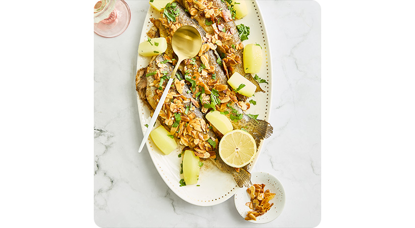  Ganze gebratene Fische auf Platte, mit Mandelbutter, Petersilie und Kartoffeln angerichtet, dazu Zitronenscheibe und kleine Schale mit gerösteten Mandeln.