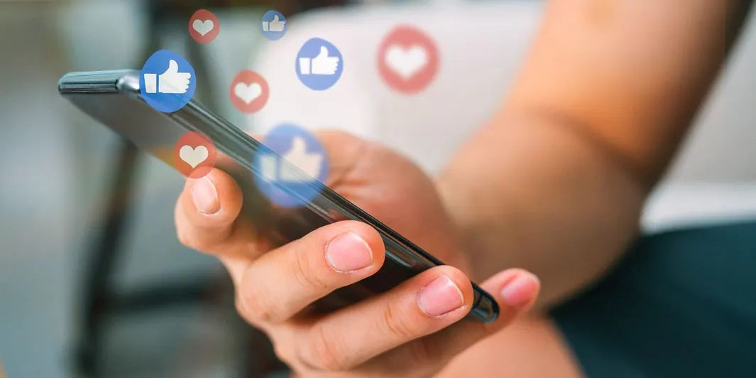 Haende halten Smartphone waehrend Like- und Herz-Symbole ueber dem Display schwebe