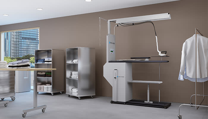 Bügel- und Finishbereich einer Gewerbewäscherei mit einem stehenden Schulthess-Bügeltisch/Presssystem, Kleiderstangen mit aufgehängten Hemden und rollbaren Wäschebehältern.