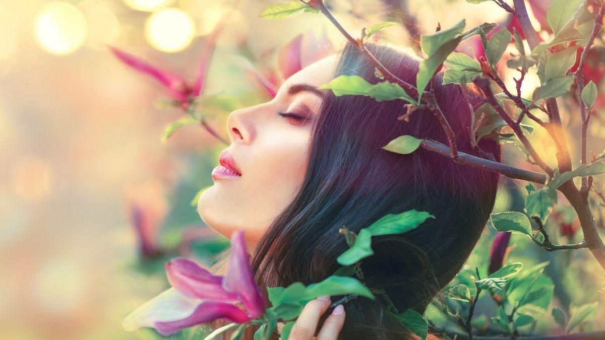Eine Person mit geschlossenen Augen, Kopf nach oben gerichtet, zwischen grünen Blättern und lila Magnolienblüten. Heller Bokeh-Hintergrund.