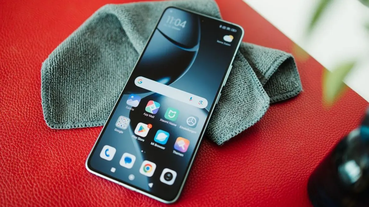 Schwarzes Smartphone mit hellem Display, Uhrzeit 11:04 und App-Symbolen auf roter, genarbter Oberfläche. Graues Tuch.