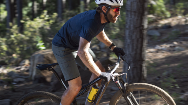 Ein Mann fährt mit einem Fahrrad auf einem Weg. Er trägt einen Helm, eine dunkle Hose und ein blaues Hemd. Bäume im Hintergrund.