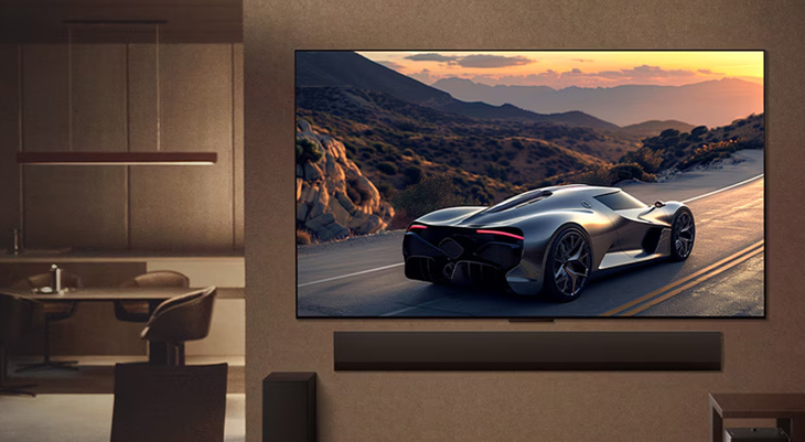 Großer, wandhängender Flachbildfernseher in modernem Wohnzimmer zeigt einen silbernen Sportwagen, der bei Sonnenuntergang auf einer Gebirgsstraße fährt; darunter ist eine schmale Soundbar montiert.