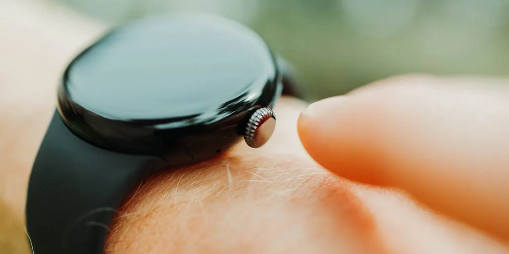 Nahaufnahme einer dunklen Smartwatch mit glänzendem Display und schwarzem Armband am Handgelenk. Ein Finger nähert sich der Krone vor unscharfem Hintergrund.