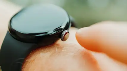 Nahaufnahme einer dunklen Smartwatch mit glänzendem Display und schwarzem Armband am Handgelenk. Ein Finger nähert sich der Krone vor unscharfem Hintergrund.