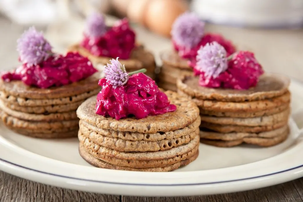 Mehrere kleine Blinis mit pinker Creme und essbaren Blüten dekoriert.